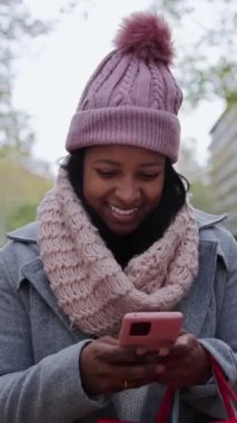 Dikey gülümseyen Afro-Amerikan kadın şehir caddesinde yürürken akıllı telefon kullanıyor. Bir kadın, yılbaşı günü bahçe bahçesinde cep telefonunu ararken eğleniyor.. 