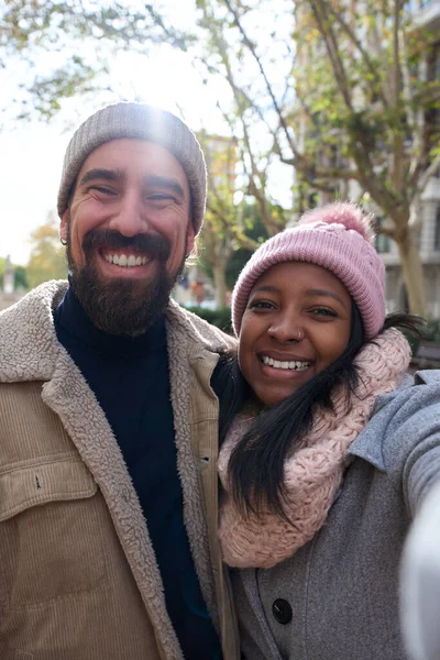 Vertical. Winter portrait of happy young tourist couple taking selfie outdoors in a city. Smiling multi-ethnic people traveling together on vacation. Cheerful man and woman posing for photo with phone