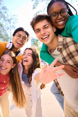 Vertical multiracial group of teenagers taking a selfie looking front camera laughing and having fun piggybacking together outdoor. Young students enjoying their free time with friends in street city clipart