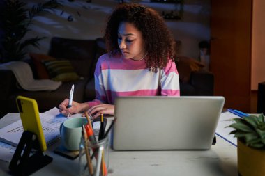 Genç Latin kadın gece evde not defterine not alarak konsantre oluyor. Afro kız işe dizüstü bilgisayarla odaklanmış. Üniversite öğrencisi oturma odasında masada oturuyor. Uzaktan çalışan genç bir kadın.