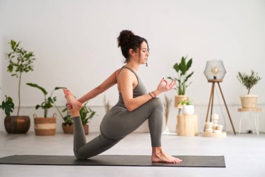 Gri spor giyimli, yoga yapan beyaz bir kızın yan görüntüsü. Tek bacaklı Kral Güvercin pozu ya da Eka Pada Rajakapotasana. Sakin bir kadın evde gözleri kapalı meditasyon yapıyor..