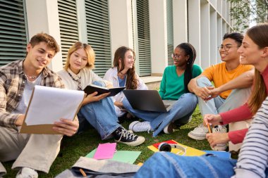 Group of young international university students sitting on the grass outside the faculty building. Friends gathered happily studying together with workbooks and laptop on campus clipart