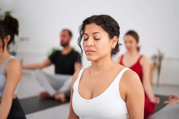 Close-up young Latina woman with eyes closed practising yoga exercises indoors. In the background a group of people meditating and concentrating in class. Healthy lifestyle in community and wellness.