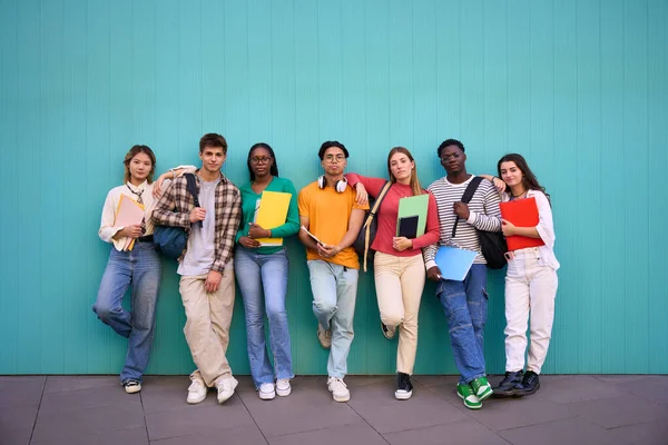 Serious international students posing standing looking at the camera. Young people multicultural friends concentrated and embracing with backpacks and workbooks in a blue wall background