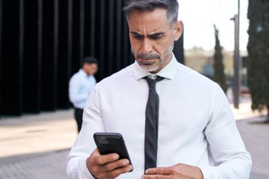 Çekici, olgun, gri saçlı iş adamı odaklanmış olmayan bir cep telefonu sokağı gibi görünüyor. Ofis dışında ciddi resmi personel. İş yerinde cep telefonu kullanan profesyonel erkek.
