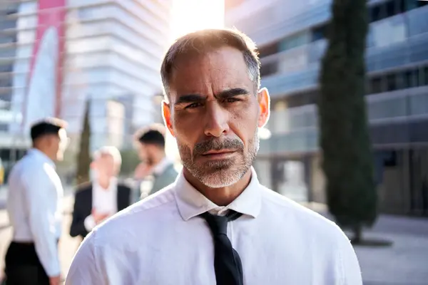Portrait of attractive middle-aged business man looks at camera with serious expression standing outdoor with unfocused colleagues in background. Confident formal professional outside office building