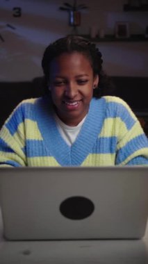 Dikey Afrikalı kız, akşam odasında laptoptan video görüşmesi yapıyor. Öndeki kadın konuşuyor ve gece evde web kamerasını selamlıyor. Genç bir bayan masada oturmuş internette sohbet ediyor. Genel bağlantıyı kavra.