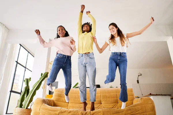 Group of multicultural excited women jumping off sofa at home together. Three girlfriends having fun enjoying private party in living room. Cheerful people in community and active relationships.