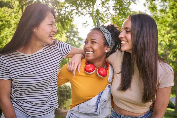 Group of young multicultural nice women laughing enjoying standing together in park on sunny day. Three females smile excited while having fun outdoors. Generation z, lifestyle, friendship relations.