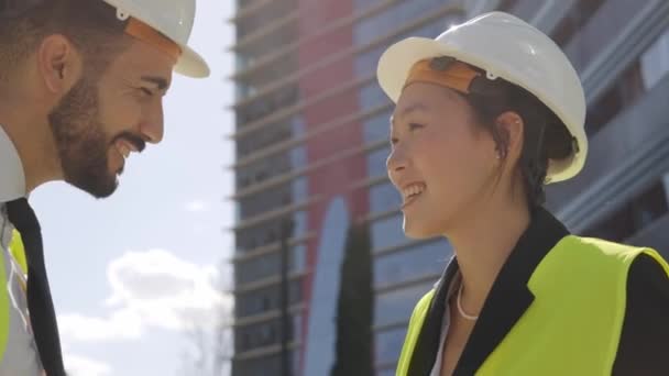 屋外で丁寧にチャットする2人の技術者 多種多様な建築家が新しいプロジェクトについて笑顔で話しています イエローベスト 安全ヘルメットのプロの女性男性のクローズアップ — ストック動画