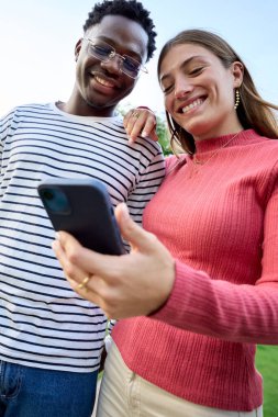 Dikey. Genç, çok kültürlü bir çift, gülümseyip, ellerinde akıllı telefonlara bakıyorlar. Bahçede duruyorlar. Dışarıdaki cep telefonunu kullanan neşeli ve mutlu insanlar. Beyaz kız ve Afrikalı adam.