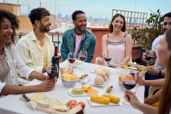 Happy group of multiracial young adult friends enjoying lunch together outdoors. Cheerful people gathered smiling with glass of red wine on summer day celebrating a birthday snack party on rooftop