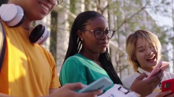 陽気な若い多人種大学の学生のグループは 彼らのモバイルセルに没頭し テクノロジーは大学のキャンパスの外に座っている友人を中毒しました スマートフォンを使った笑顔の遺伝子Zの人々 — ストック動画