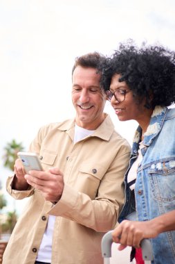 Dikey odaklanmış olgun turist çifti ayakta gülümsüyor ve ellerinde cep telefonuyla dışarıda bekliyorlar. Güneşli günlerinde yer arıyor ve tatillerinin keyfini çıkarıyorlar.