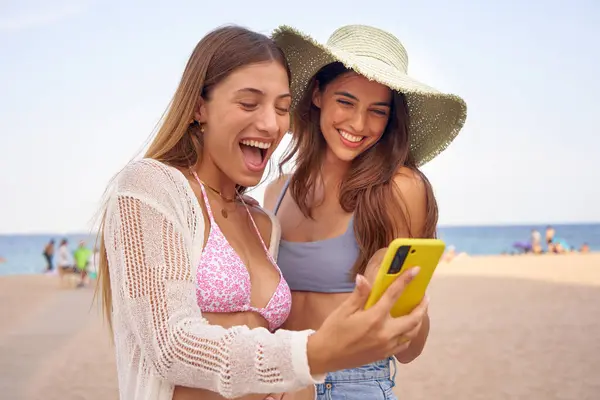 Two girl friends sharing a mobile device to check social media on the beach. Young women check smartphone and laugh a lot. They smile together while watching videos on the phone.