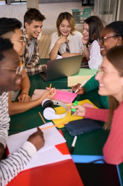 Kampüsteki kafeteryada kız arkadaşlarıyla birlikte dizüstü bilgisayar kullanan gülümseyen genç bir üniversite öğrencisinin dikey görüntüsü. Fakülte binasında birlikte eğitim gören çok ırklı akademisyenler