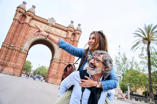 Cheerful mature tourist attractive couple pointing at something on city street. Man with grey hair gives piggyback ride to smiling woman. Happy adult people in love enjoying sightseeing on vacation