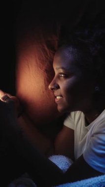 Flört uygulaması kullanan dikey genç Afrikalı kız. Yatak odasındaki karanlıkta uzanmış, cep telefonuyla sohbet eden kadın. Gülümseyen ve teknolojiye bağımlı mutlu bir kadın. Z geninde sosyal ortam kaydırılıyor