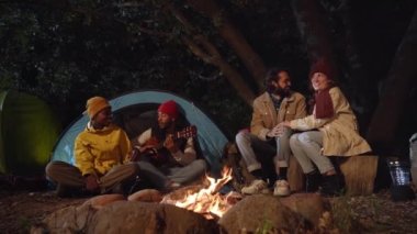Group of friends with guitar near bonfire and camping tent outdoors overnight . Multiracial colleagues charismatic sitting beside the fire stake playing on the guitar discussing and singing.