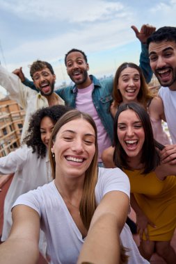 Dikey genç bayan çatıda neşeli arkadaşlarıyla grup selfie 'si çekiyor. Neşeli insanlar poz veriyor. Dışarıdaki kameralara heyecanlı yüz ifadeleri takınıyorlar. Fotoğraf için mutlu kollarını kaldırıyorlar.