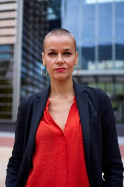 Dikey stil sahibi, modern Kafkasyalı, zarif kırmızı giyinmiş yetişkin kadın ofis alanında duruyor. Güçlü, kendine güvenen, kısa saçlı, dışarıda kameraya ciddi bakan bir kadın.