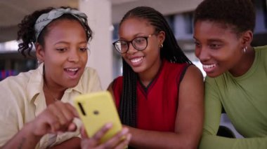Bir tanesi akıllı telefondan komik videolar izlerken gülümseyen üç güzel siyahi kadın grubu. Afrika kökenli Amerikalı kadın arkadaşlar birlikte neşeli görünürler.