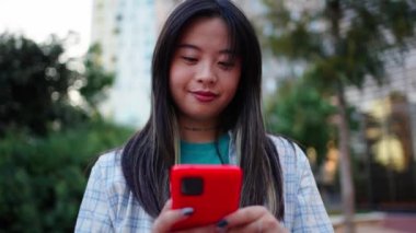 Şehir caddesinde cep telefonuyla gülen neşeli Asyalı genç kadın. Z jenerasyonu Çinli güzel kadın hareketli ekranda mutlu görünüyor sosyal medyadaki videonun içeriğinden keyif alıyor. Ağ bağımlısı insanlar 