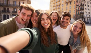 Bir grup genç insan binanın önünde selfie çekerken mutlu bir şekilde gülümsüyor ve eğleniyorlar. Boş zamanlarında keyifli bir anı paylaşıyorlar.
