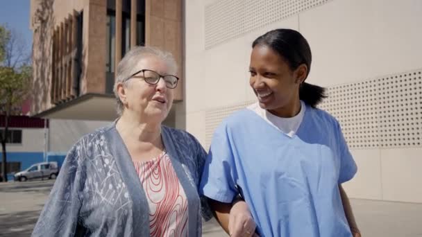 笑笑年轻的非洲裔美国人助手与灰白头发的退休祖母手挽手走在大街上 为需要日常照顾和照顾的长者提供家庭帮助 养恤金领取者的伴侣 — 图库视频影像