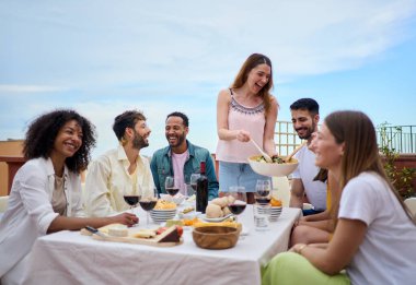 Bir grup insan bir masanın etrafında toplanıyor, yemek ve şarabı paylaşırken aynı zamanda gülümsüyor ve eğleniyor. Ev sahibesi misafirlerine yemek servisi yapar. Bir bahar günü arkadaşlarla öğle yemeği.