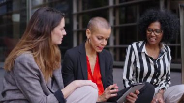 Çok kültürlü yetişkin iş kadınlarından oluşan bir grup ofis binasının dışında sessizce sohbet ediyorlar. Profesyonel kadınlar açık havada tablet bilgisayar kullanarak ve arayarak pazarlama stratejisinden bahsediyorlar.