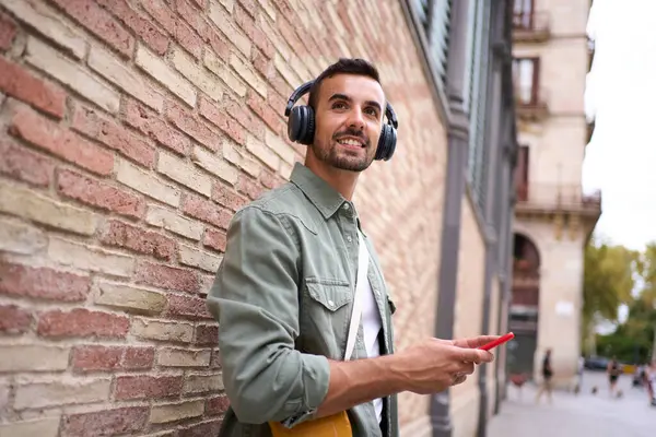 Young handsome man smiling with wireless headphones while holding cell phone leans against brick wall. His gesture suggests relaxation as he enjoys happy looking at something outdoors. Copy space