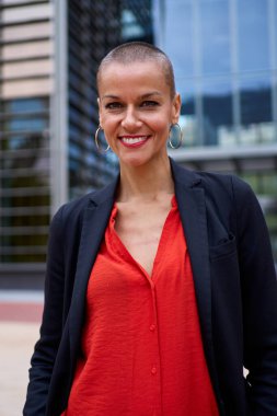 Kurumsal ofis alanında dikey şık, beyaz, şık kırmızı elbiseli yetişkin bir kadın. Güçlü ve güçlü çekici bir kadın. Kısa saçlı, gülümseyen. Açık havada kameraya mutlu bakıyor.