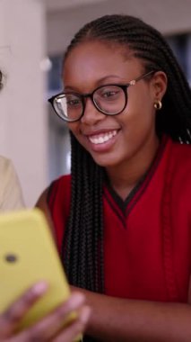 Dikey genç siyah kadınlar mutlu bir şekilde gülümsüyor akıllı telefonları tutuyor ve komik videolar izliyorlar. Z jenerasyonu Afro-Amerikan bayan arkadaşları kapalı alanda neşeli görünüyor. Merkezdeki kıza odaklan. 