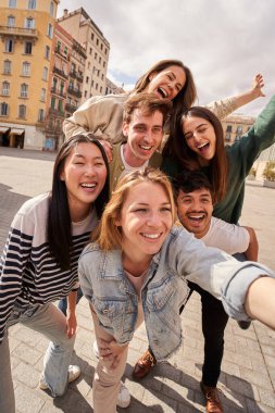 Gençlerden oluşan bir ekibin dikey fotoğrafı mutlu bir şekilde selfie çekiyor, gülümsüyor ve jestler yapıyor, turistik bir kentteki gezi etkinlikleri sırasında eğleniyorlar.