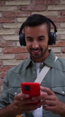 Dikey çekici, kulaklıklı, kırmızı cep telefonu kullanan, tuğla duvara dayanan genç bir adam. Milenyum Avrupalı erkek açık havada durup gülümseyerek gezmekten zevk alıyor. Teknolojiye bağımlı insanlar 
