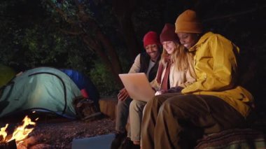 Kış gecesi kamp alanında şenlik ateşinde yan yana oturan üç gülen insan bilgisayar kullanıyor. Mutlu genç arkadaş grubu sörf ağının tadını çıkarıyor. Açık doğa kaçamağı için kamp çadırı..