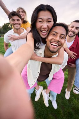 Dikey. Çok ırklı bir grup genç arkadaş parkta sırtüstü selfie çekiyorlar ve çiftler tarafından kucaklanıyorlar. Mutlu milenyum insanları dışarıda kameraya bakarak birlikte gülümsüyor. Üniversite öğrencileri