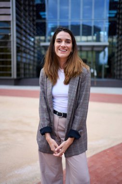 Dikey şık yetişkin işi. Takım elbiseli beyaz kadın şirket ofisinde duruyor. Başarılı bir kadın, el ele tutuşmuş gülümseyerek poz veriyor ve dışarıda kameraya mutlu bakıyor. Bina arkaplanı