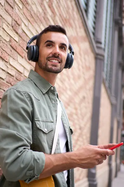 Vertical young handsome man smiling with wireless headphones holding cell phone leans against brick wall. His gesture suggests relaxation as he enjoys happy looking at something outdoors. Copy space