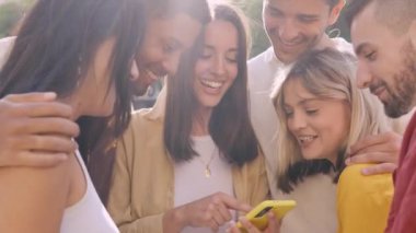 Genç bir grup insan dışarıda sosyal medyayı izlerken cep telefonuyla eğleniyor. Akıllı telefon uygulamasındaki içerikten hoşlanan neşeli öğrenci topluluğu. Genç çok ırklı arkadaş grubu 