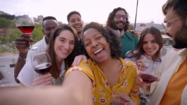 POV Mutlu genç çok ırklı insanlar kameraya bakıyor sosyal medyada arkadaşlarıyla çatı terasında bir etkinliği paylaştıkları için selfie çekiyorlar, dans ediyorlar, eğleniyorlar ve şarap kadehleri tutuyorlar.
