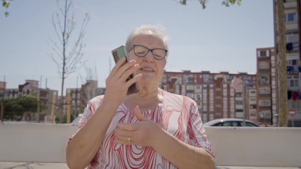 快乐的白种人老妇人 戴着眼镜坐在户外用手机聊天 放宽老年女性接听亲戚打来的电话 使用技术装置享受夏日晴天的成年人 — 图库视频影像