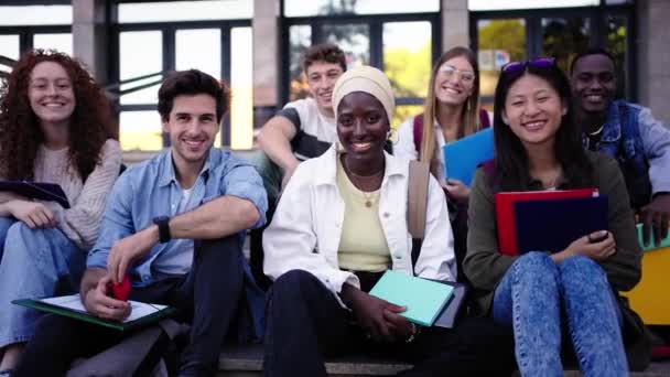 多族裔群体微笑着 学生们坐在楼梯上 手握折叠式的文件夹 凝视着大学楼外的摄像头 校园内多文化年轻人同班同学的快乐生活理念 — 图库视频影像