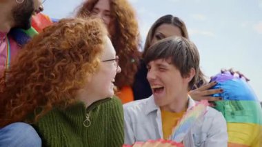 Çeşitli gençlerden oluşan bir grup balonlarla ve gökkuşağı bayraklarıyla gururla kutlama yapıyor ve mavi gökyüzü altında LGBTQ topluluğuna destek veriyorlar.