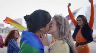 İki genç kadın, açık havada bir LGBT etkinliğinde gökkuşağı bayrağı taşırken birbirlerini şefkatle öperler. Onların jestleri aşkı ve eşitliği temsil ediyor, seyirciler arasında gülümseme ve heyecana neden oluyor.
