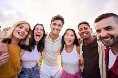 Bir grup mutlu çok ırklı arkadaş parkta dışarıda eğleniyor. Çeşitli neşeli genç insanlar ayakta durup kameraya bakıyorlar ve hep birlikte gülümsüyorlar. Sarılıp gülüyorlar.