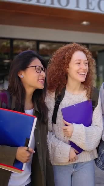 垂直的 一群快乐的多种族青年学生背着文件夹和背包离开了图书馆大楼 开玩笑 笑着走路 大学校园中的社区 — 图库视频影像