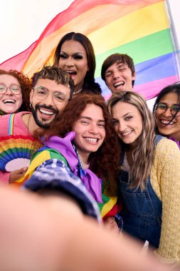 Dikey. Bir grup genç arkadaş selfie çekiyorlar ve ellerinde gökkuşağı bayrağı tutarak eşcinsellerin gurur gününü kutluyorlar. LGBT halkı gülümsüyor ve dışarıda eğleniyor.
