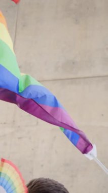 Dikey. LGBT gökkuşağı bir duvarın önünde rüzgarda sallanıyor. El ele tutuşan gay gurur bayrağı. Çeşitli insanlar dans ediyor ve bayrak altında eğleniyor. Lezbiyen, gey, biseksüel, homoseksüel, transseksüel sosyal hareketler